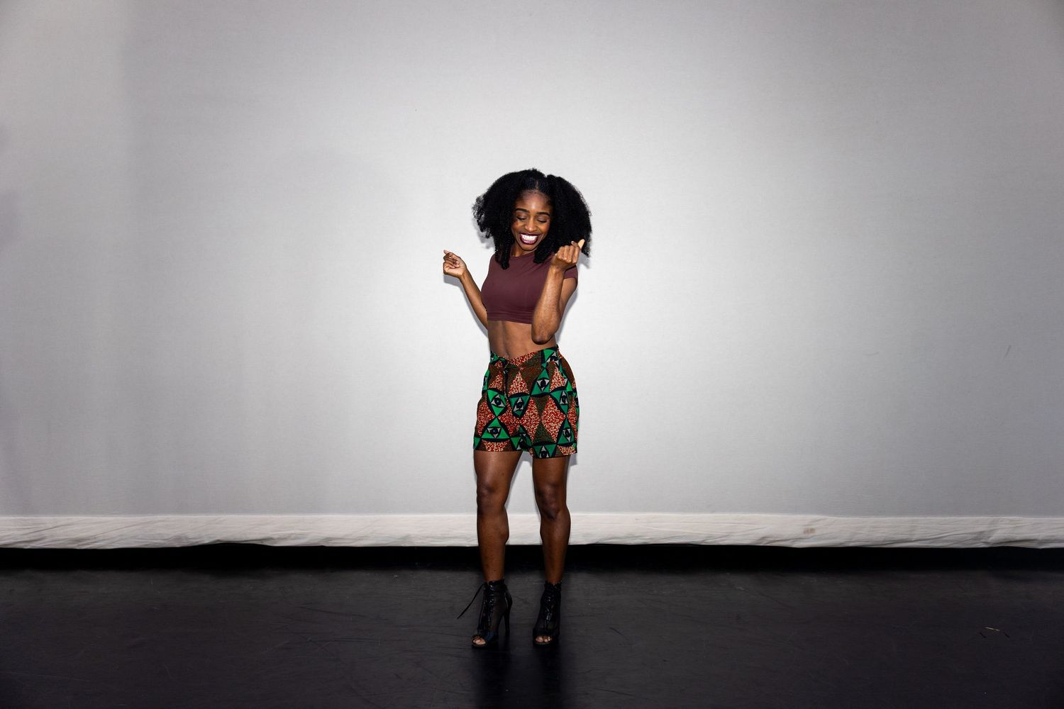 Woman in colorful African print skirt and purple crop top poses joyfully in a studio.