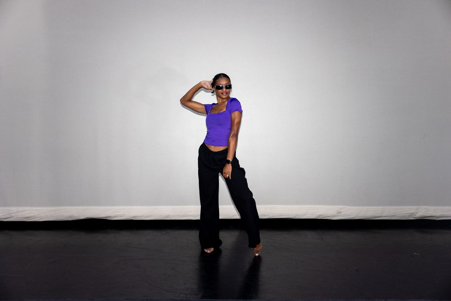Woman in purple top and black pants posing confidently against a white wall on a dark floor.