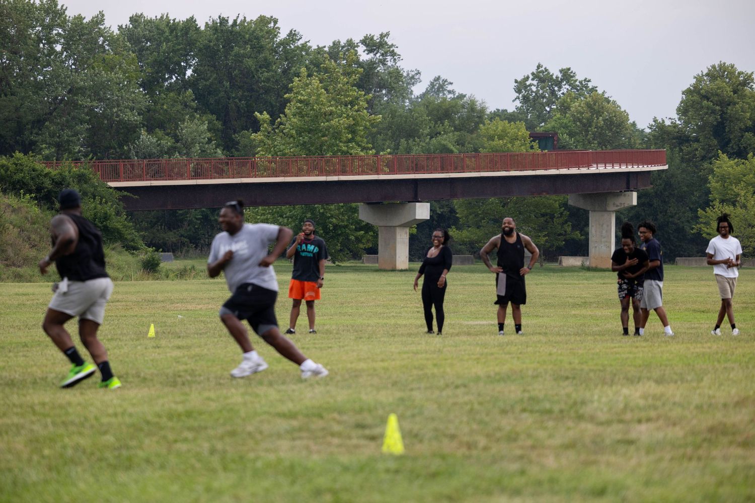 Group of young athletes practicing football drills on a green field near a bridge on a summer day.