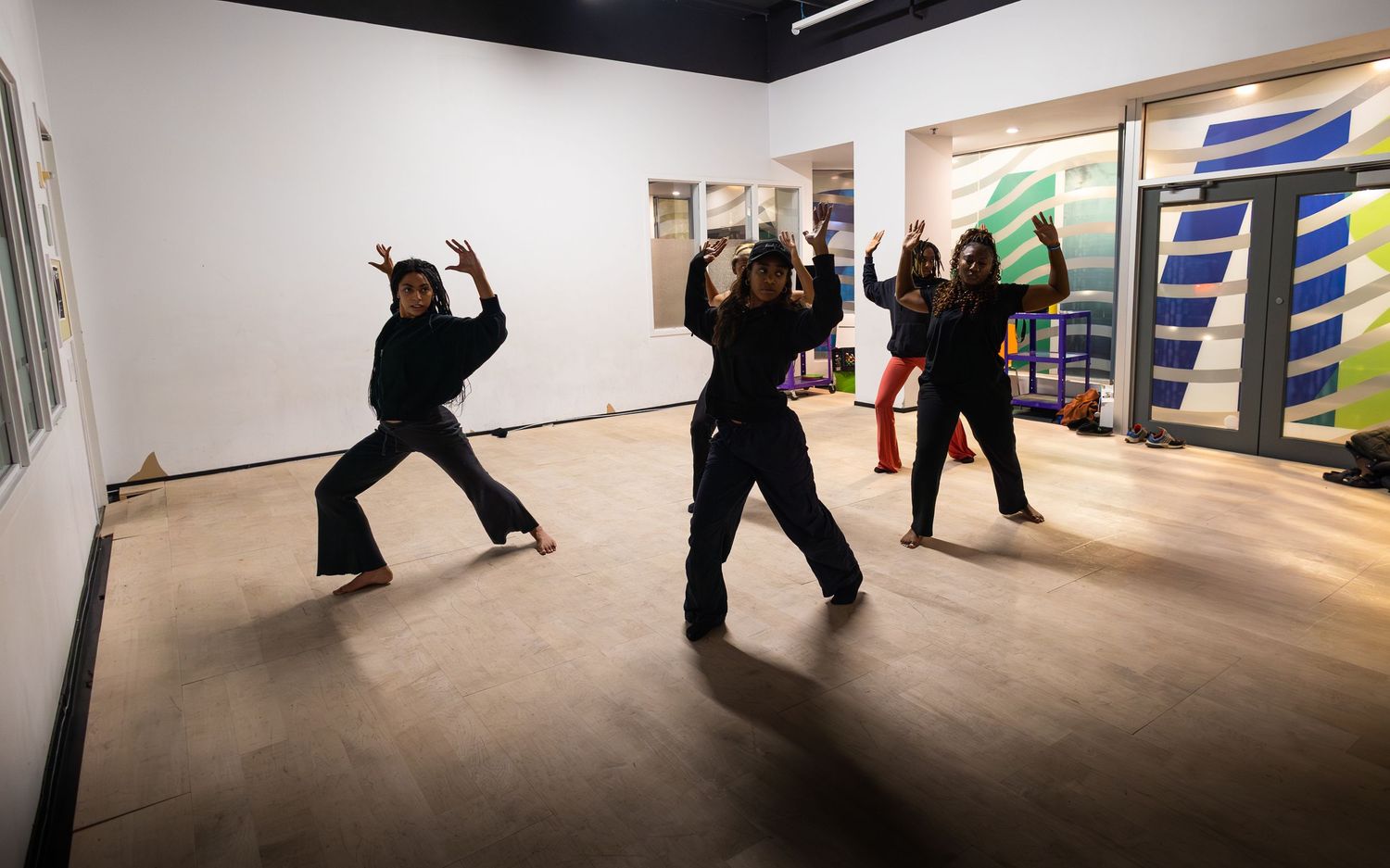 Group of dancers in black attire performing expressive moves in a bright white gallery space with colorful artwork.