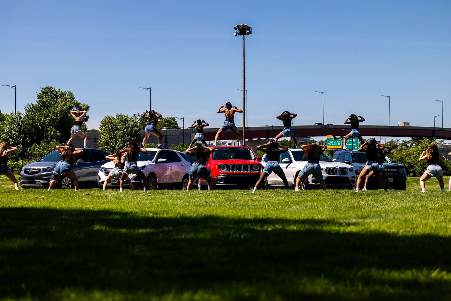 Group of dancers performing outdoor routine on grass near parked cars on a sunny day.