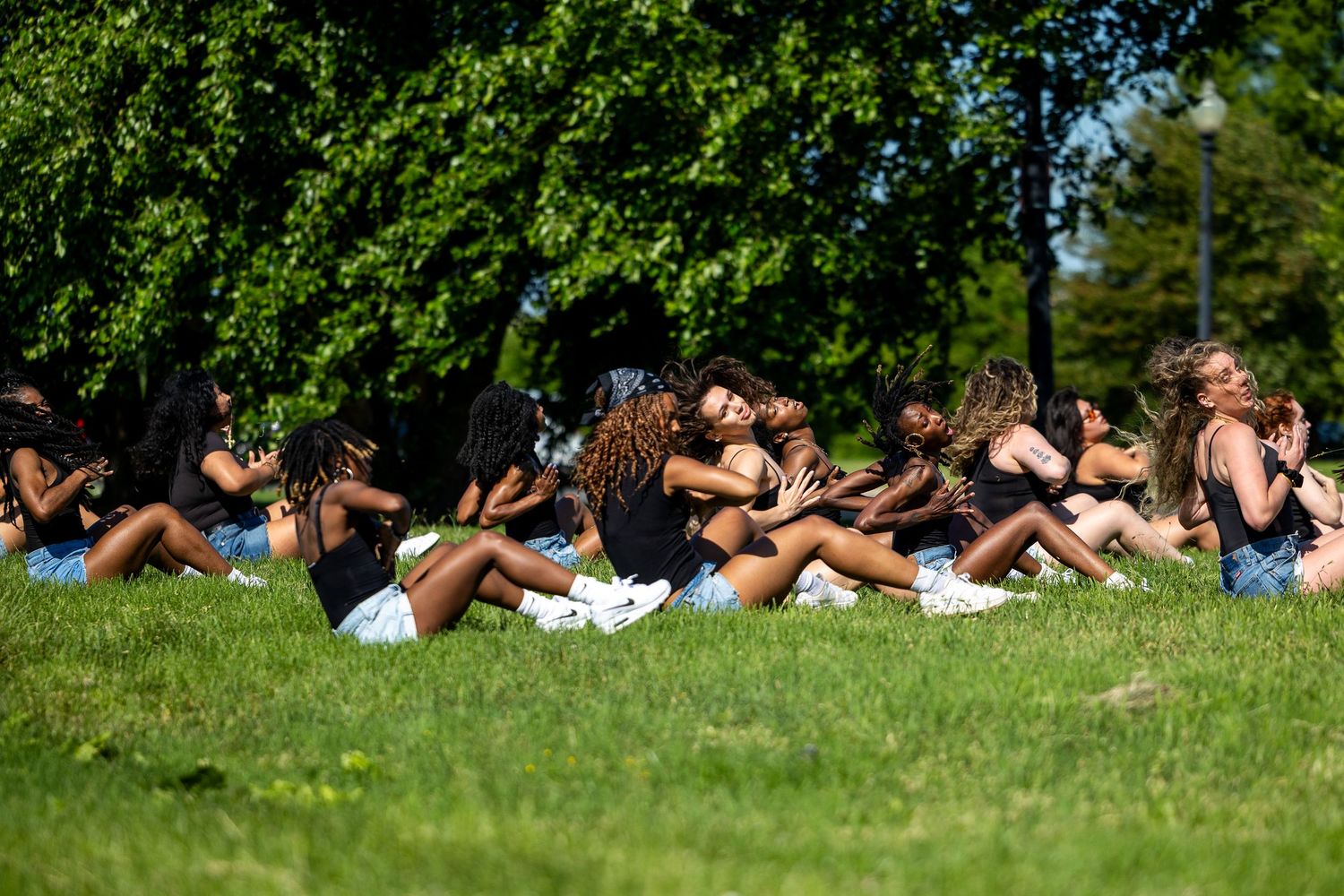A large group of young women sitting together on green grass in the shade of trees on a bright sunny day.