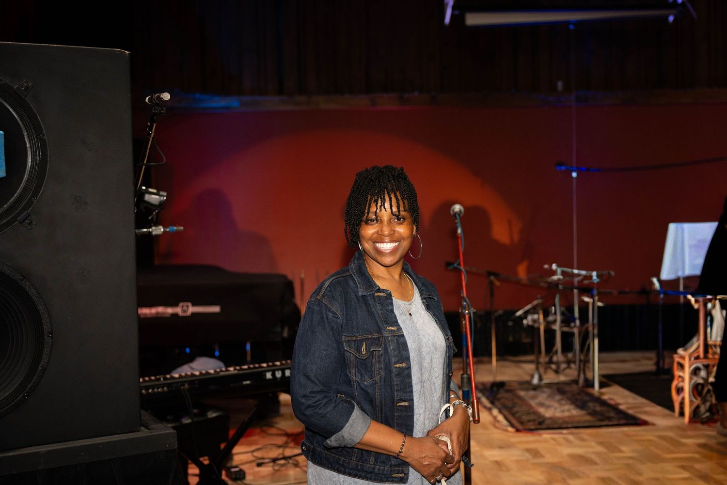A smiling woman in a denim jacket stands in a recording studio with microphones and instruments in the background.