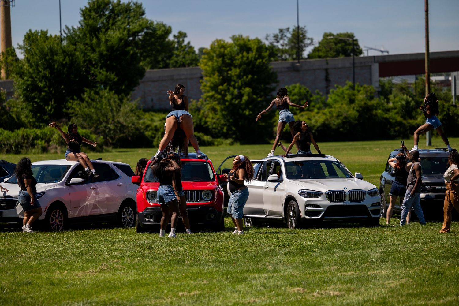 Cheerleaders performing stunts on top of cars parked on a grassy field during an outdoor practice session.