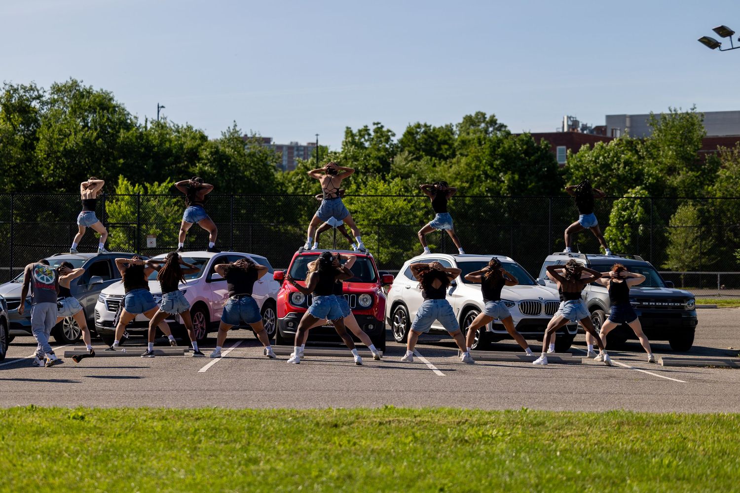 A cheerleading squad performs synchronized stunts in a parking lot surrounded by cars and green trees.