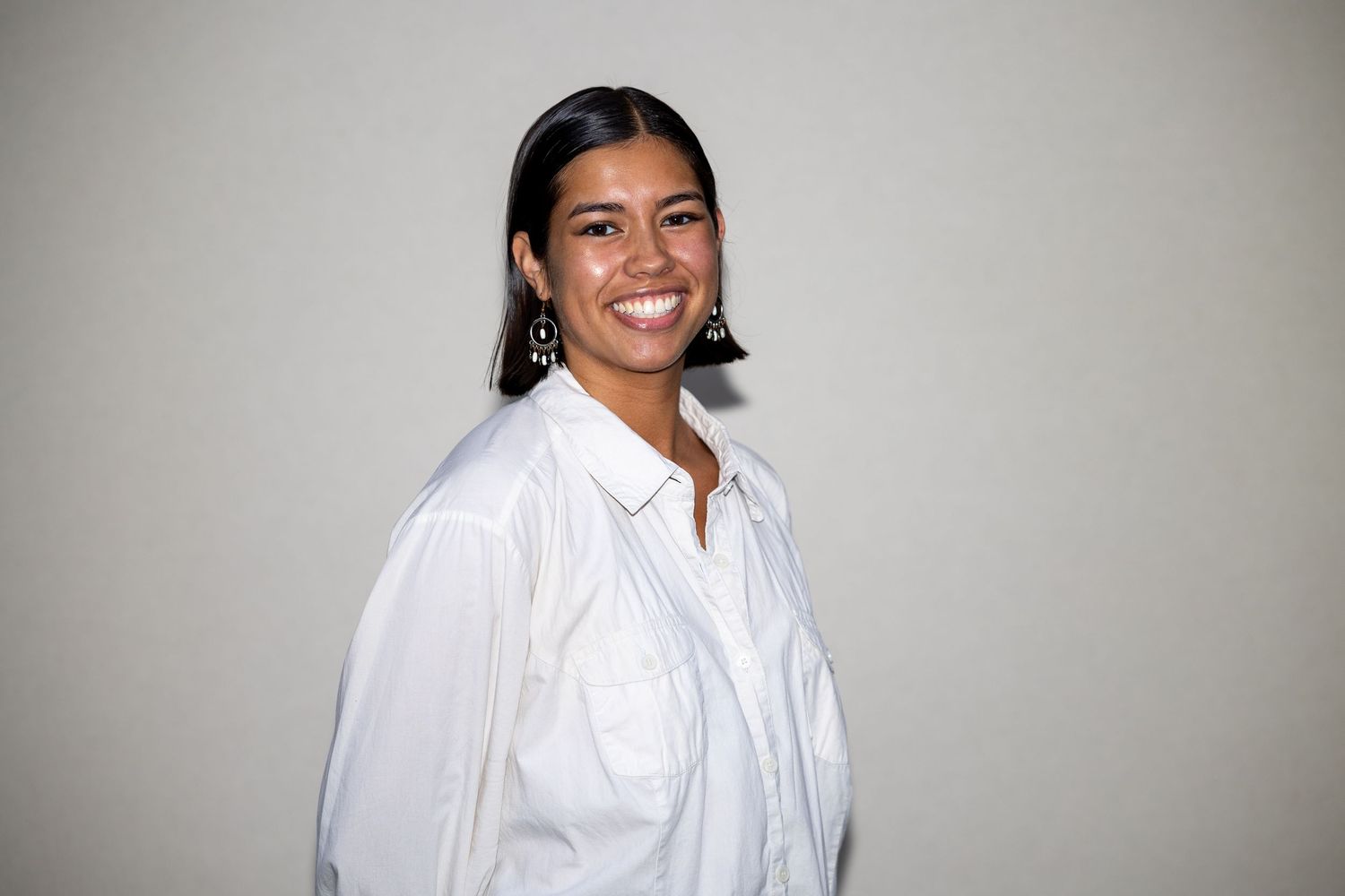 A smiling woman in a white button-down shirt with dark drop earrings poses against a light gray background.