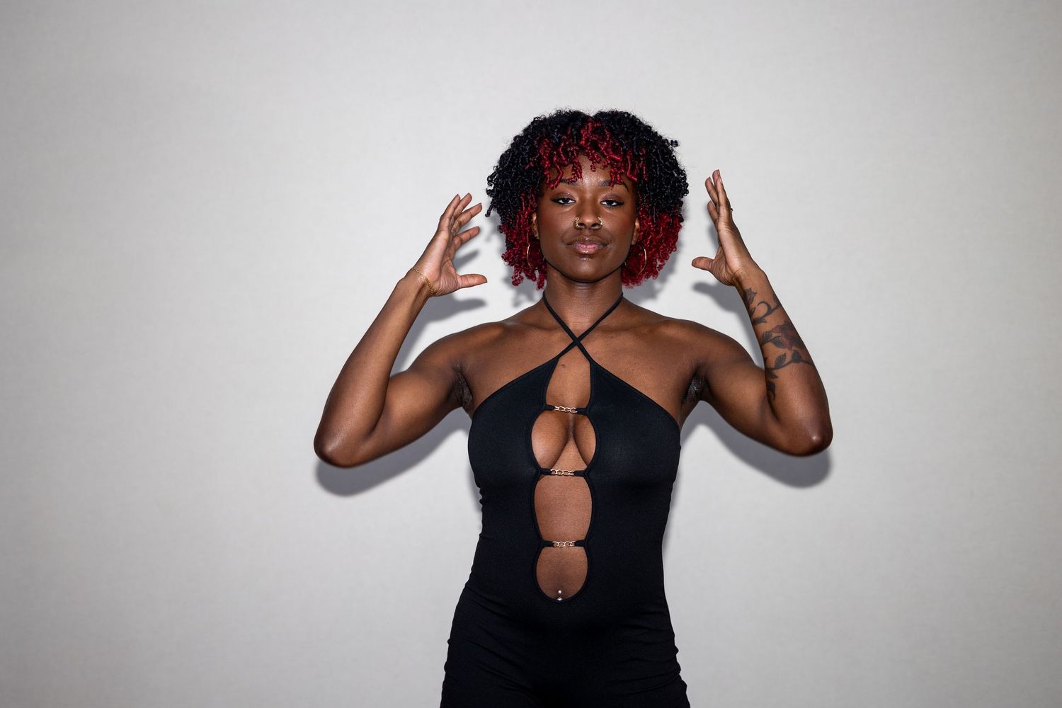 Woman with red curly hair in a black cutout dress, posing with raised hands against a white background.