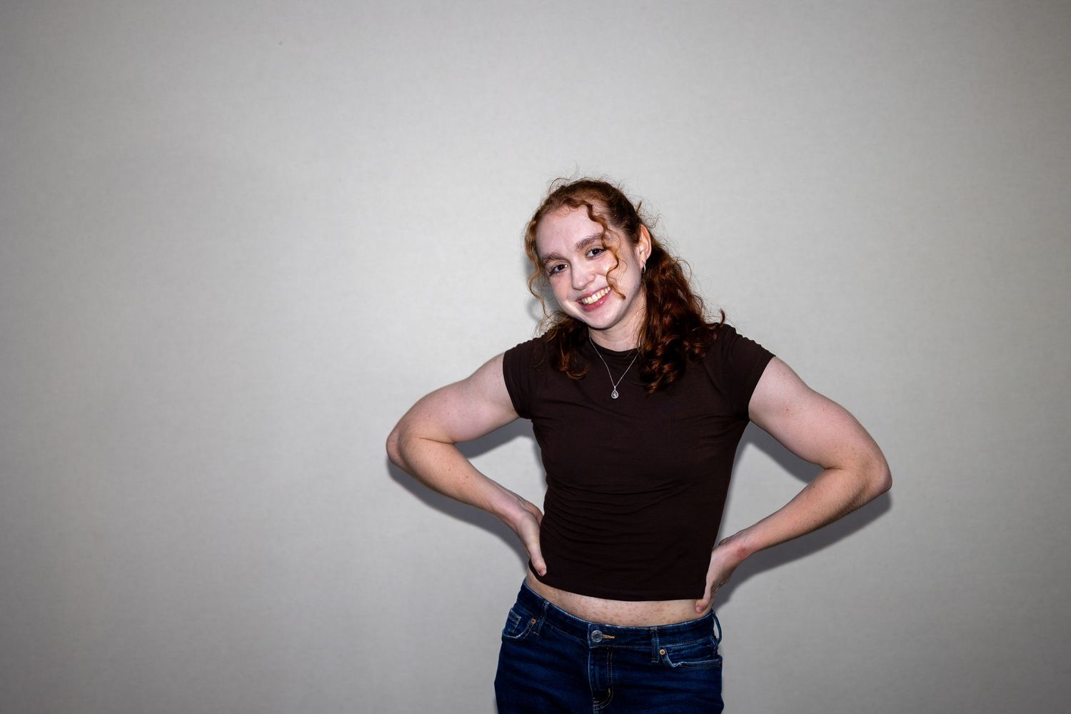 Smiling redhead woman in brown crop top and jeans poses with hands on hips.