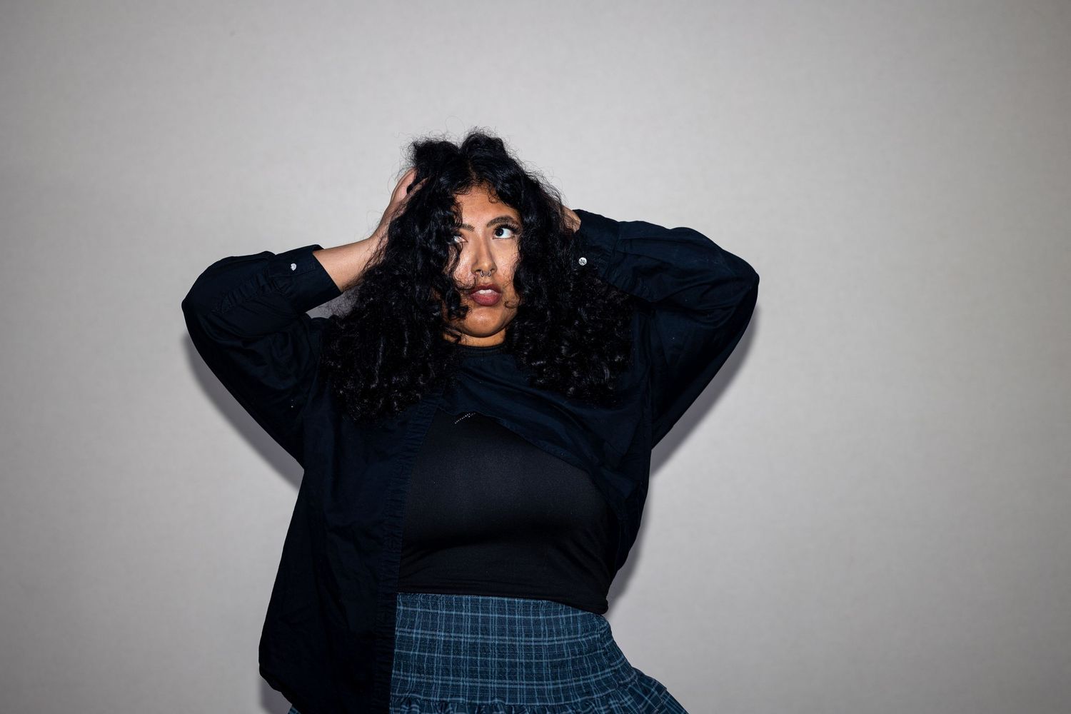 Young woman with curly hair in navy sweater and plaid skirt poses dramatically with hands in hair.