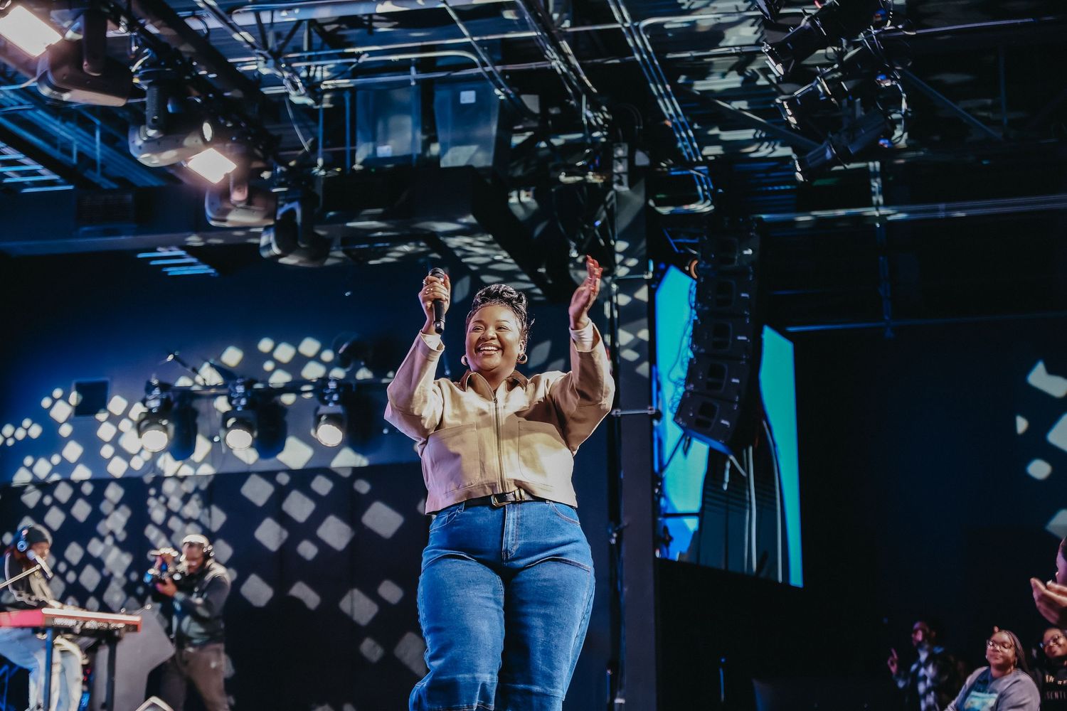 Performer in denim and beige top dances on stage with dramatic blue lighting and geometric light patterns in background.