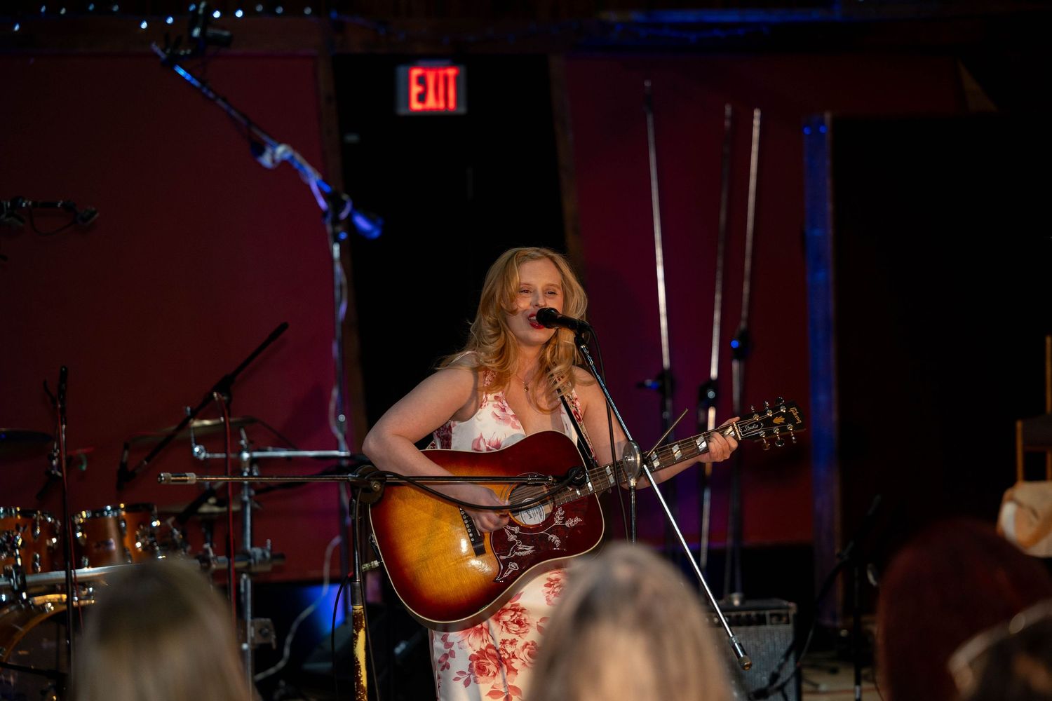 Blonde female musician performing live on stage, singing into microphone while playing acoustic guitar in dimly lit venue.