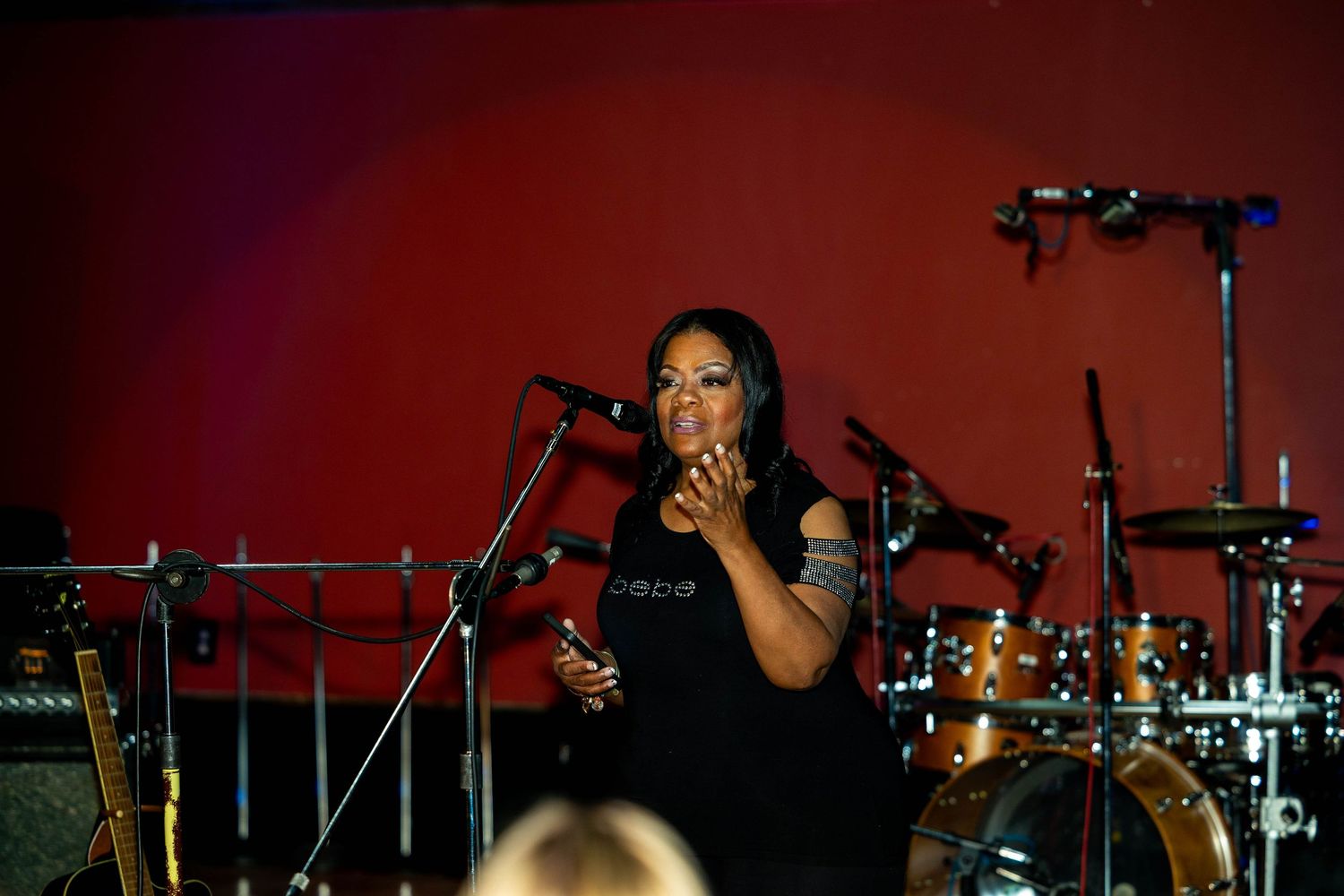 Female vocalist performing on stage in black dress with microphone stands and drum kit against red backdrop.