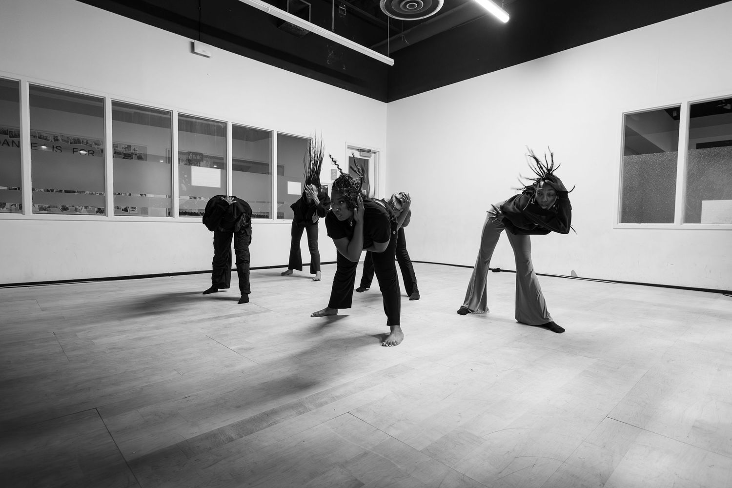 Black and white photo of dancers in dark costumes bowing dramatically in a white studio space.