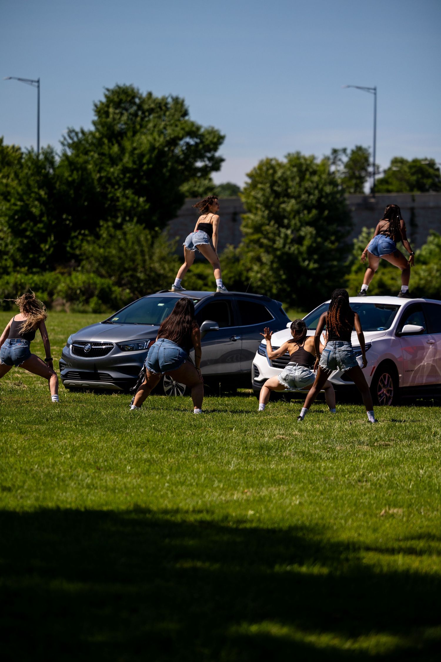 Group of young women in denim shorts dancing and posing around SUVs on a green lawn on a sunny day.