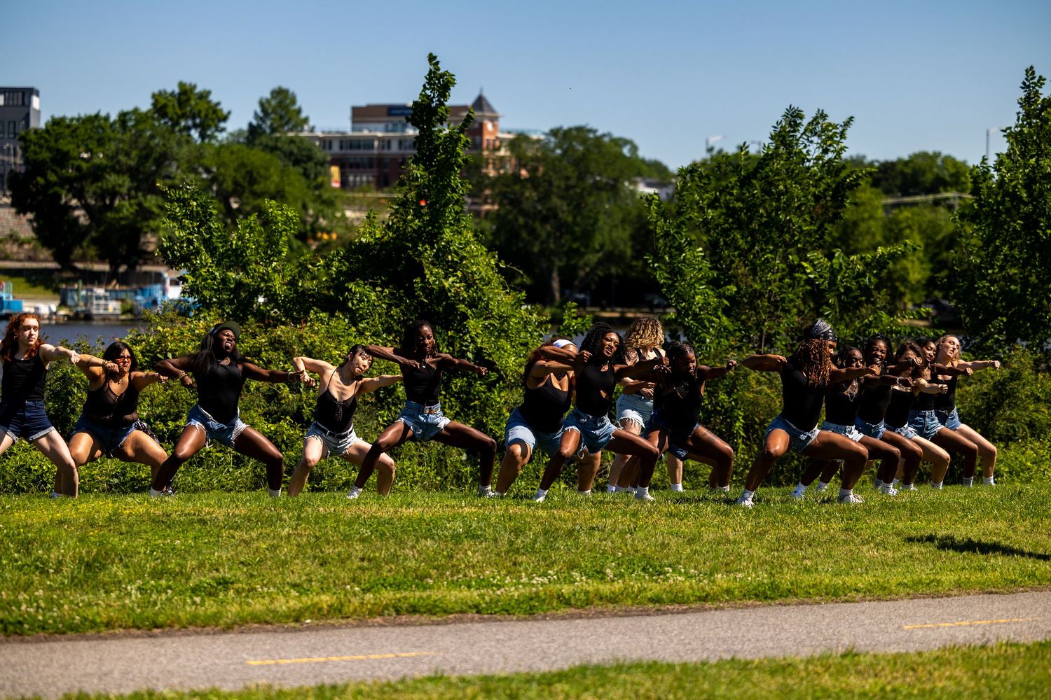 Large group of dancers performing synchronized jumps outdoors in a park with lush green trees on a sunny day.