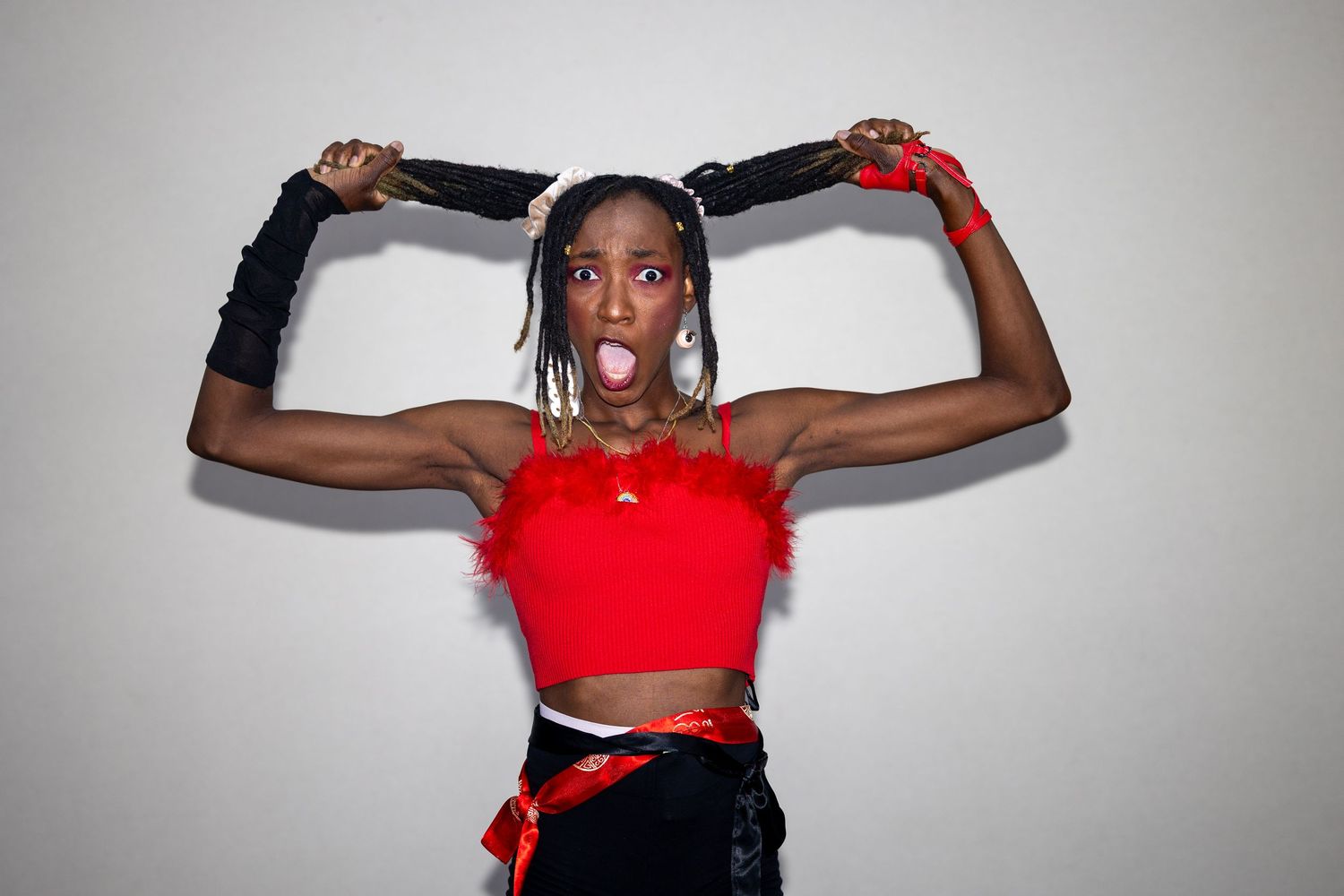 Woman in red crop top with feathers, black gloves, holding braids up, making a fun expression against gray background.
