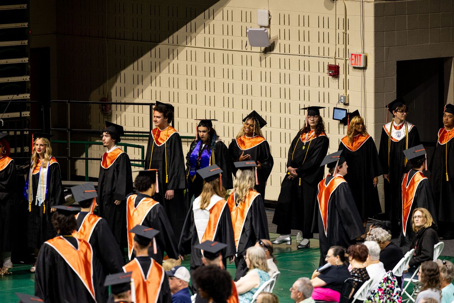 Graduates in black robes and orange sashes stand during a graduation ceremony inside a large indoor venue.