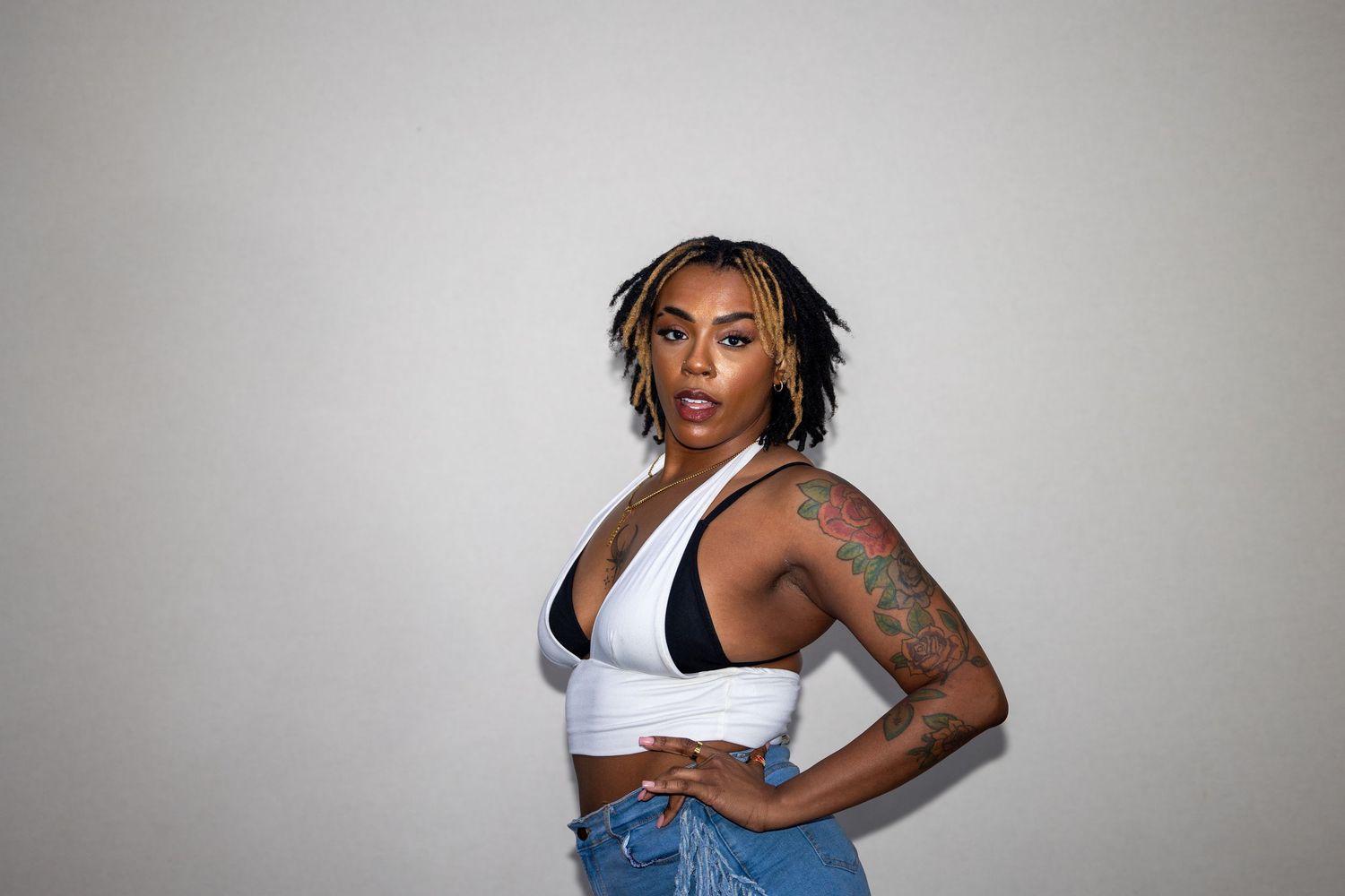 A confident woman in a white crop top and jeans with a tattoo poses with hands on hips against a white wall.