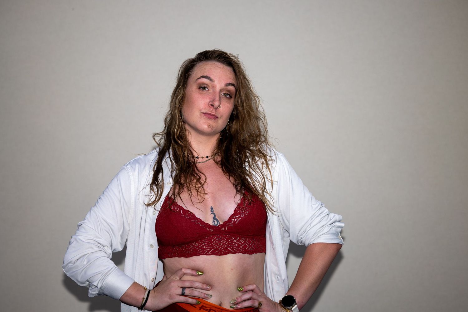 A woman in a red lace bralette and open white shirt poses with hands on hips against a light background.