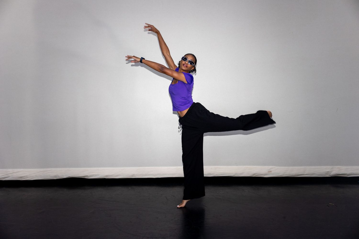 A dancer in a purple top and black pants executes a graceful leg extension in a studio.