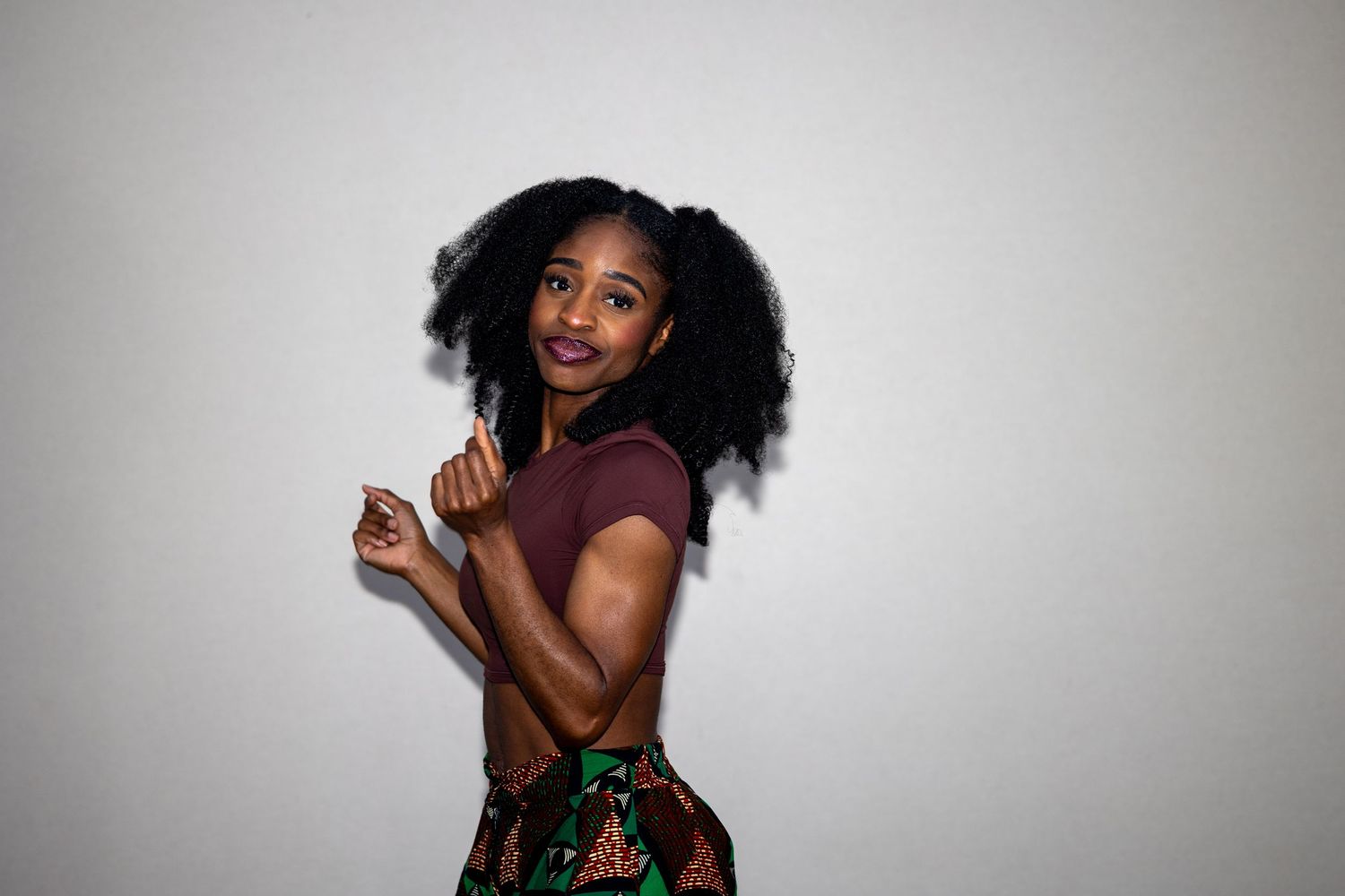 Young Black woman in maroon crop top and colorful printed skirt posing with a playful smile.