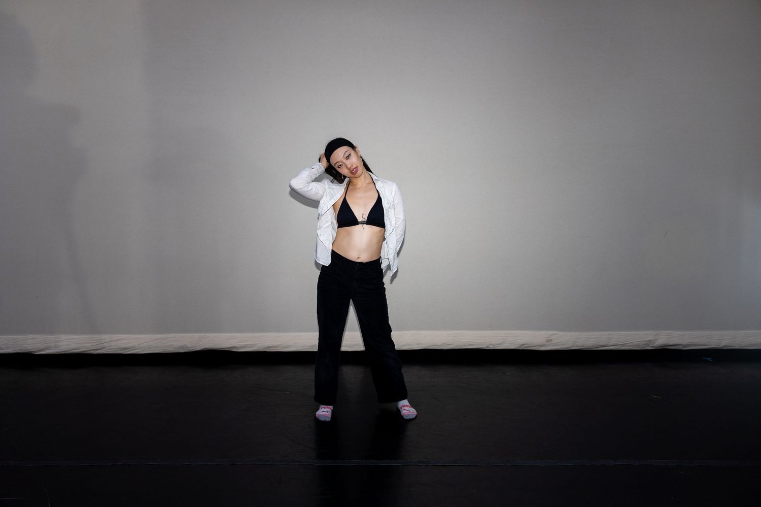 A woman in a black sports bra, black pants, and open white shirt poses barefoot on a dark studio floor.