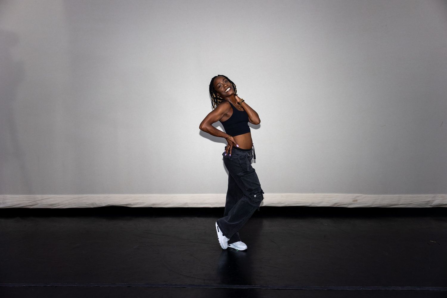 A dancer in baggy jeans and a black crop top poses casually on a dark studio floor.