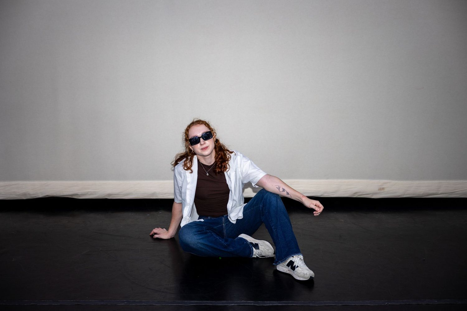 Redhead woman in sunglasses sitting casually on black floor in jeans and white shirt.