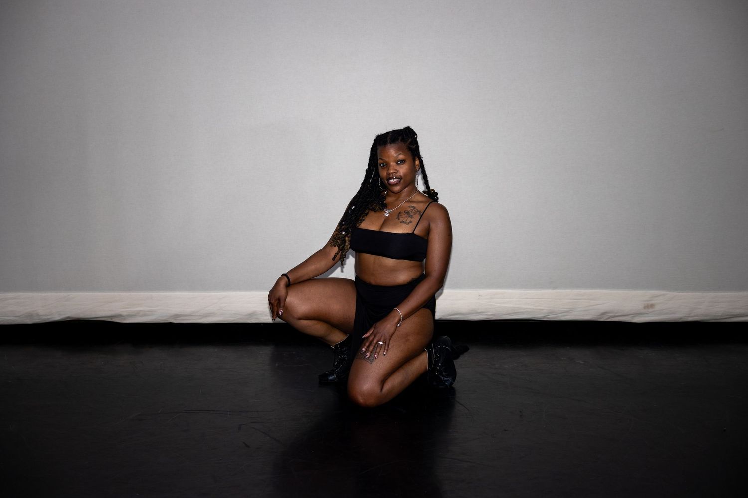 A dancer in all-black attire kneels gracefully on a dark studio floor against a gray wall.
