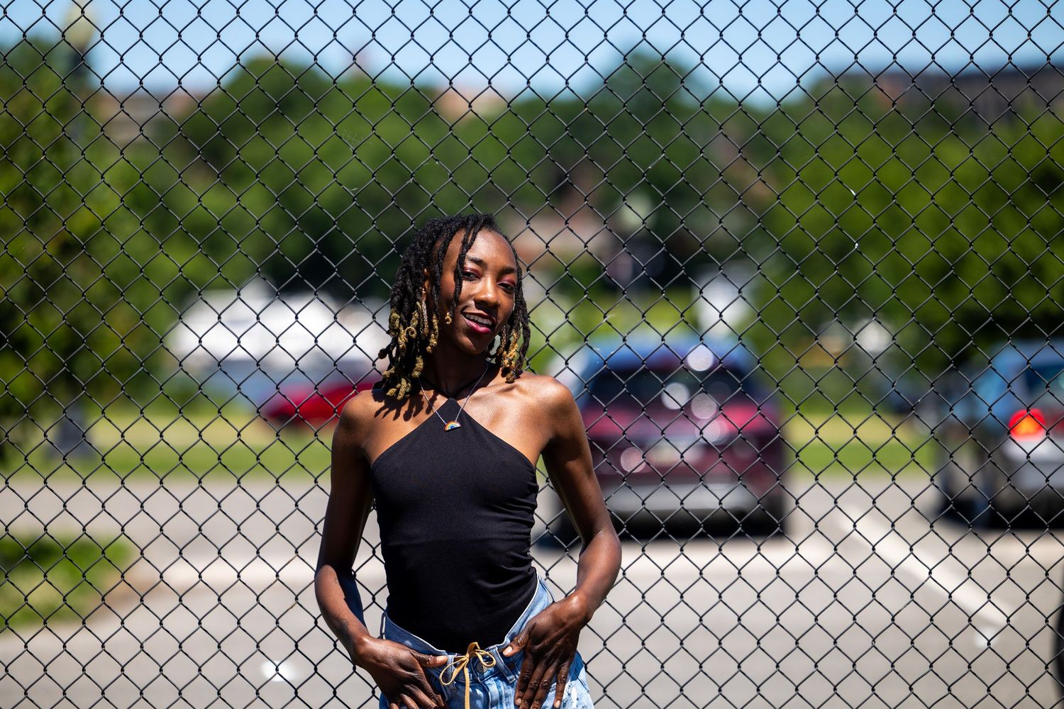 Young Black woman in black one-shoulder top poses confidently behind chain-link fence outdoors on sunny day.