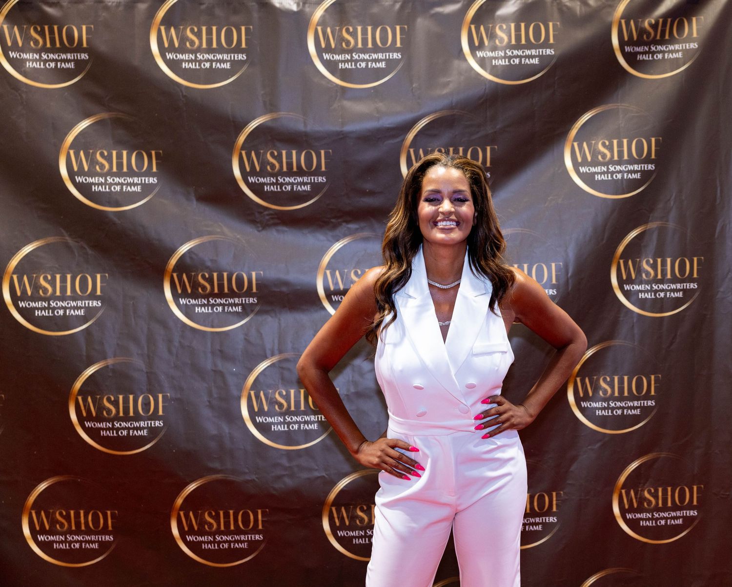 Smiling woman in white sleeveless jumpsuit posing at WSHOF Hall of Fame backdrop.