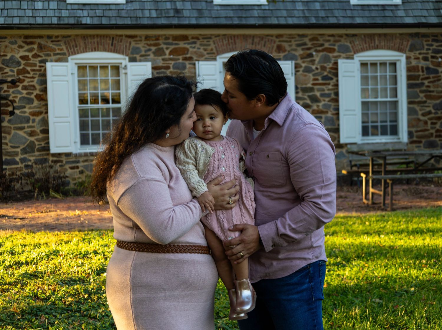 A family poses outdoors, parents kissing their baby girl in pink near a stone building at dusk.
