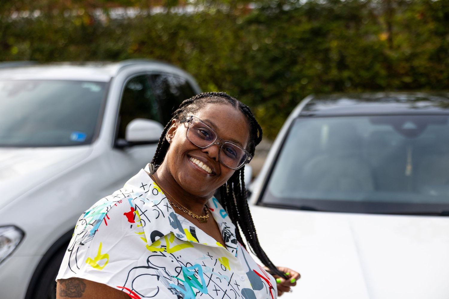 Smiling woman with braids and glasses poses outdoors near white cars on a sunny day.