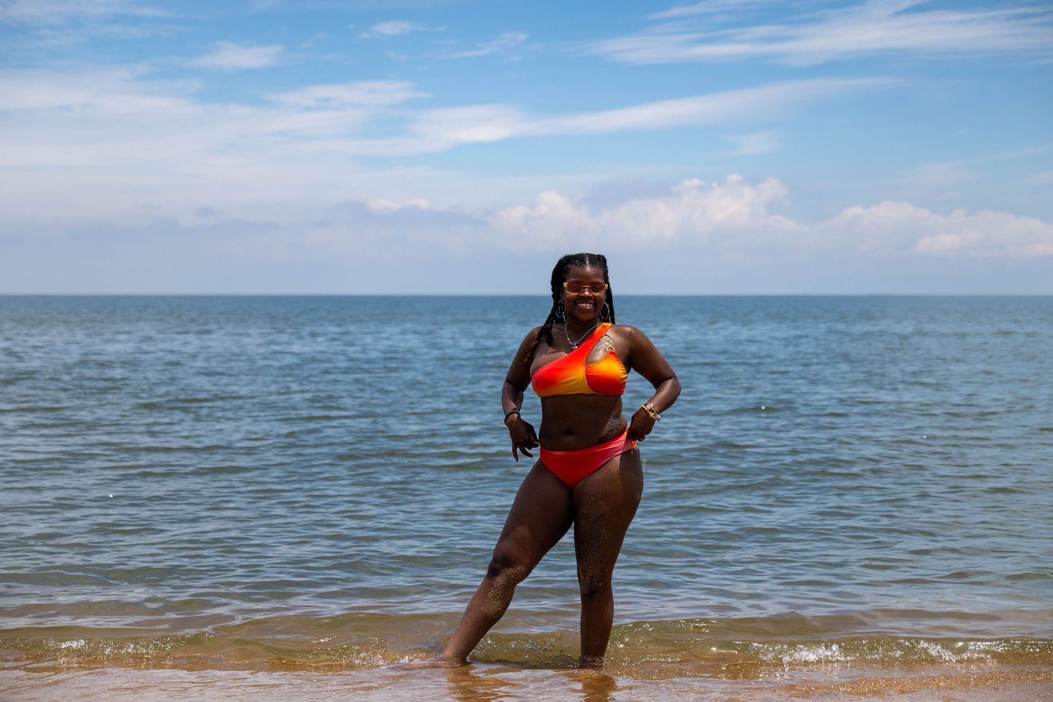 Woman in colorful bikini standing on sandy beach with calm ocean waves and blue sky behind her.