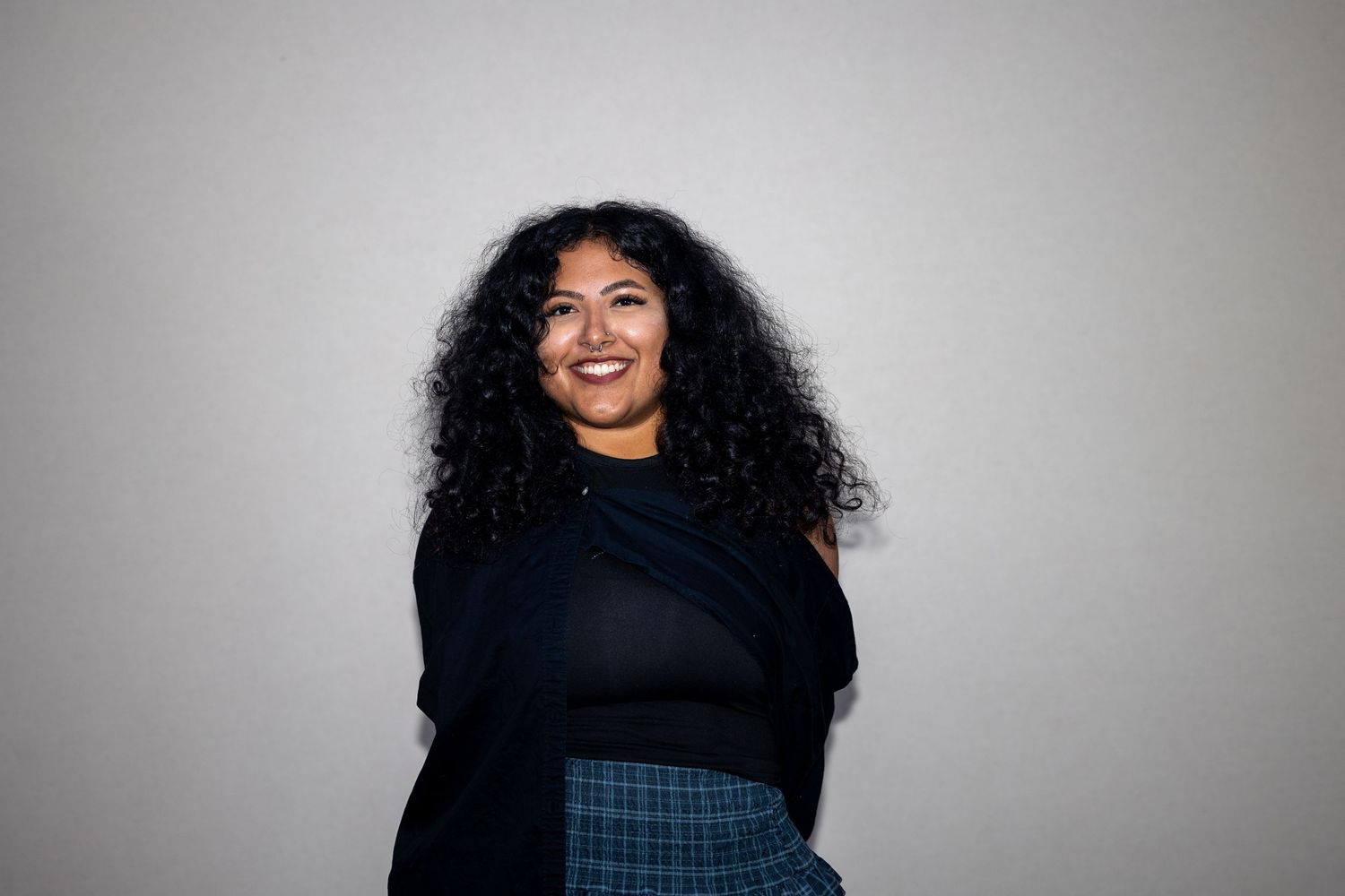 Young woman with curly hair smiling confidently in a dark top and plaid skirt against a light background.