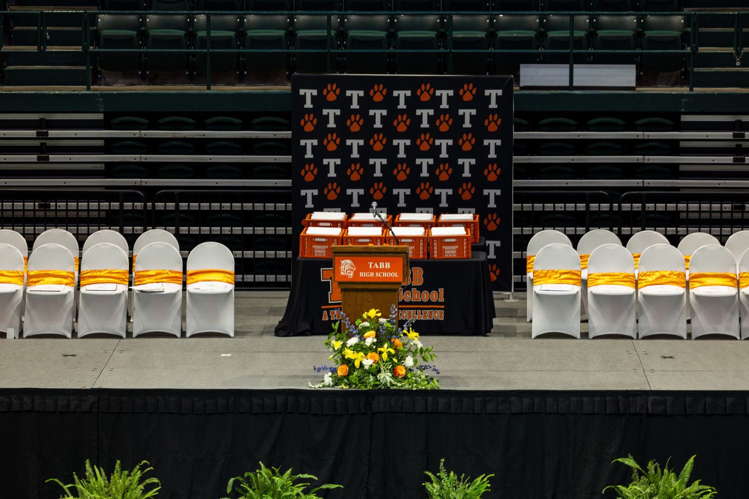 Empty graduation stage with podium, orange and white chairs, and floral arrangements in an arena.