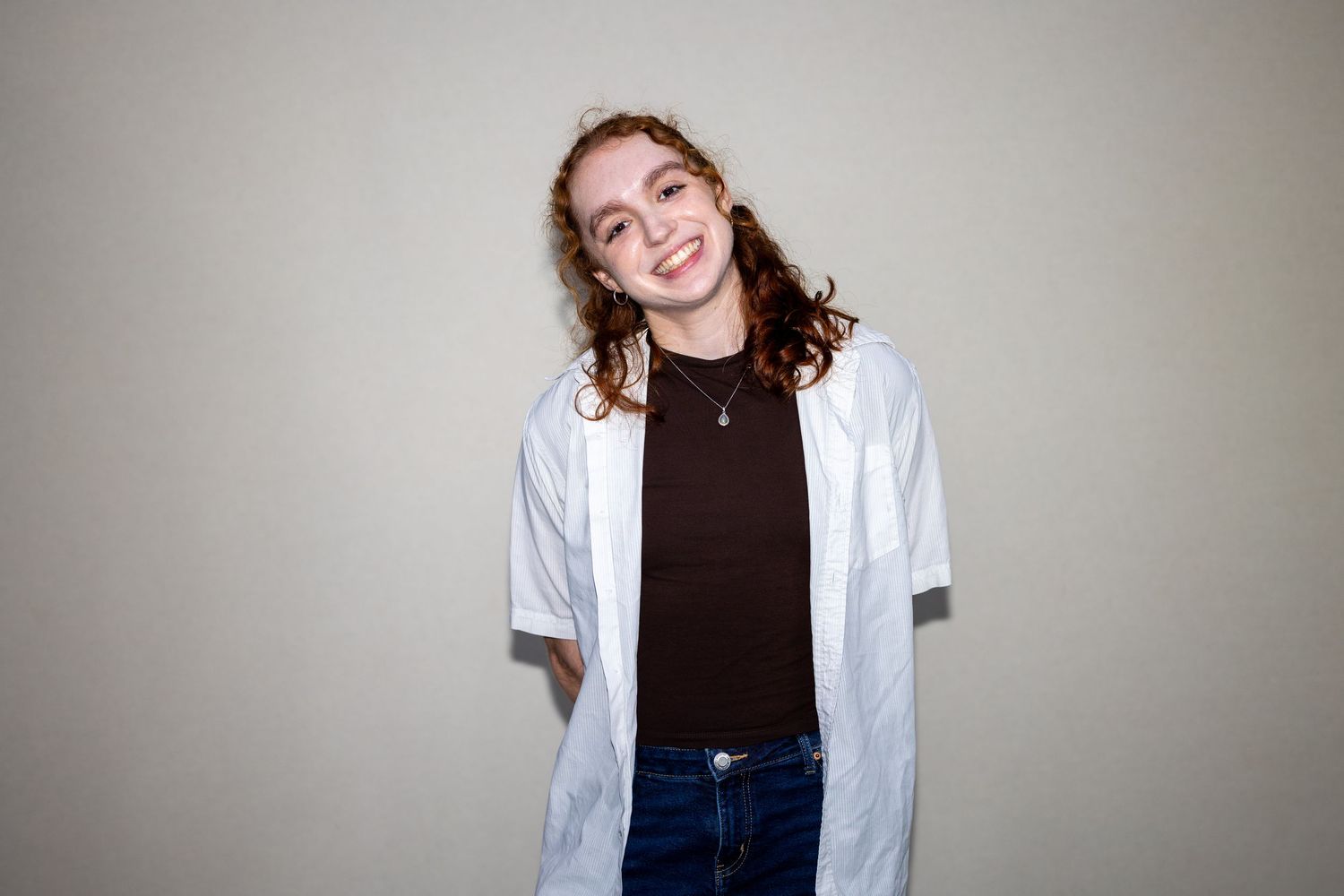 Young woman with curly red hair smiling in a white shirt over a dark top and jeans.