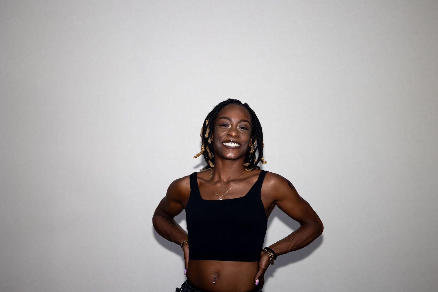 A cheerful Black woman with braids smiles broadly in a black crop top with hands on her hips.