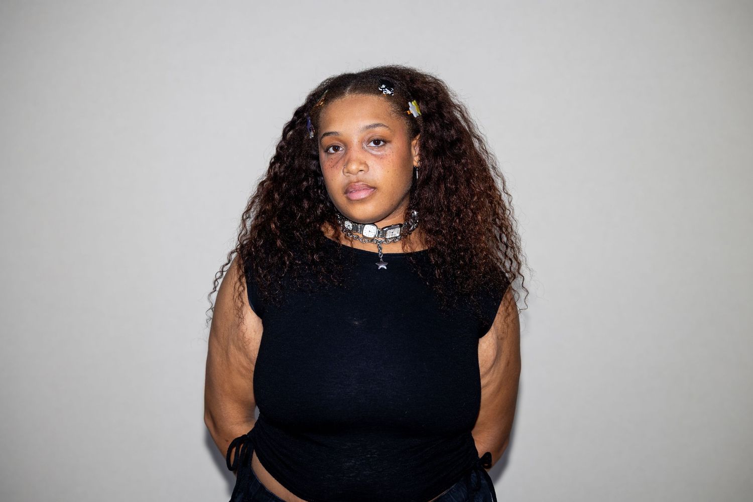 A young woman with voluminous curly hair wears a black crop top and choker necklace against a gray background.