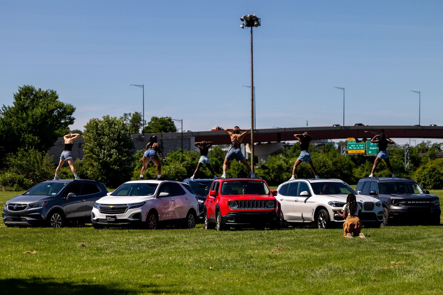 People standing on top of colorful SUVs and crossover vehicles parked on grass near a highway overpass.