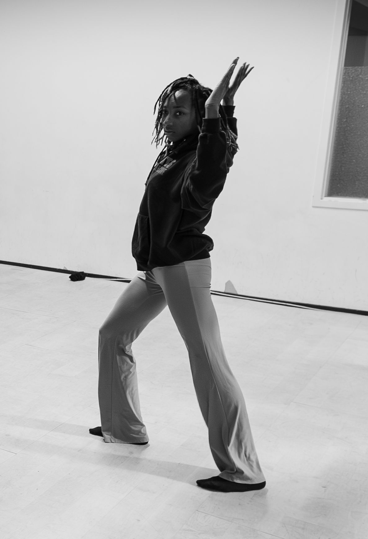 Black and white photo of a dancer in flared pants and hoodie striking a dynamic pose in a studio.