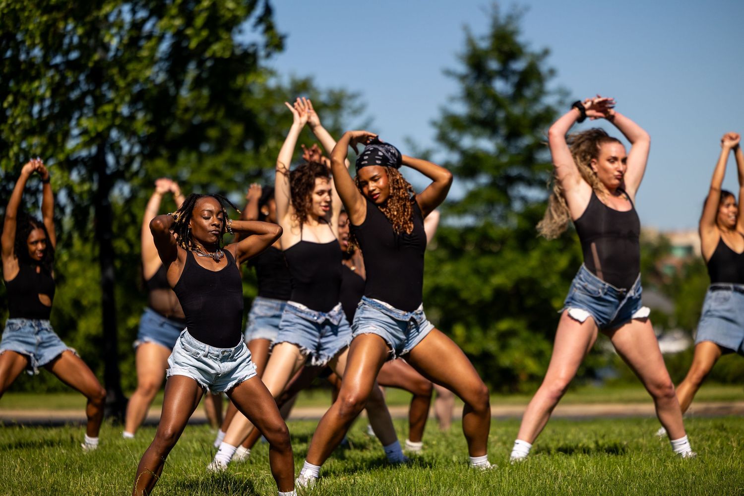Group of young women in black tops and denim shorts performing a synchronized dance routine outdoors on green grass.