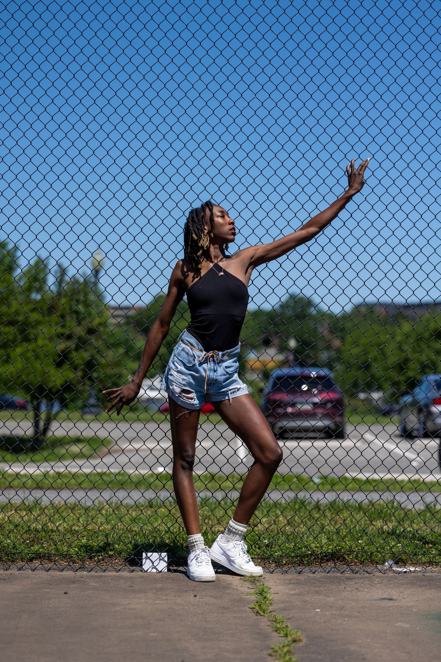 Young Black woman in black top and denim shorts poses gracefully by a chain-link fence on a sunny day.