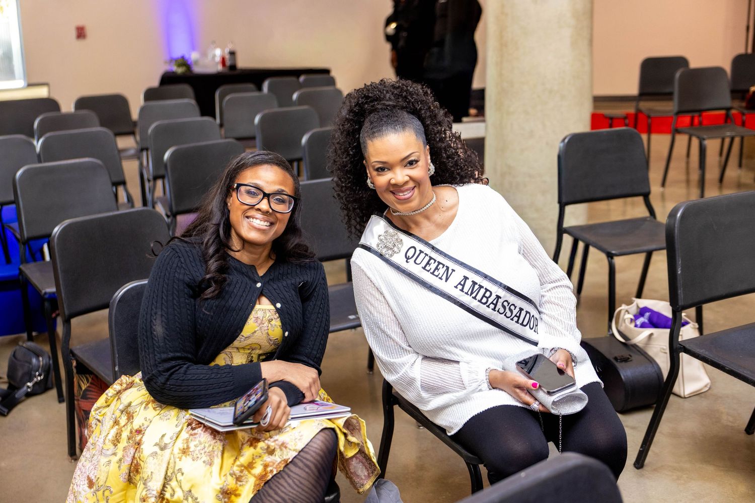 Two smiling women seated in a conference room, one wearing a Queen Ambassador sash, posing together for a photo.