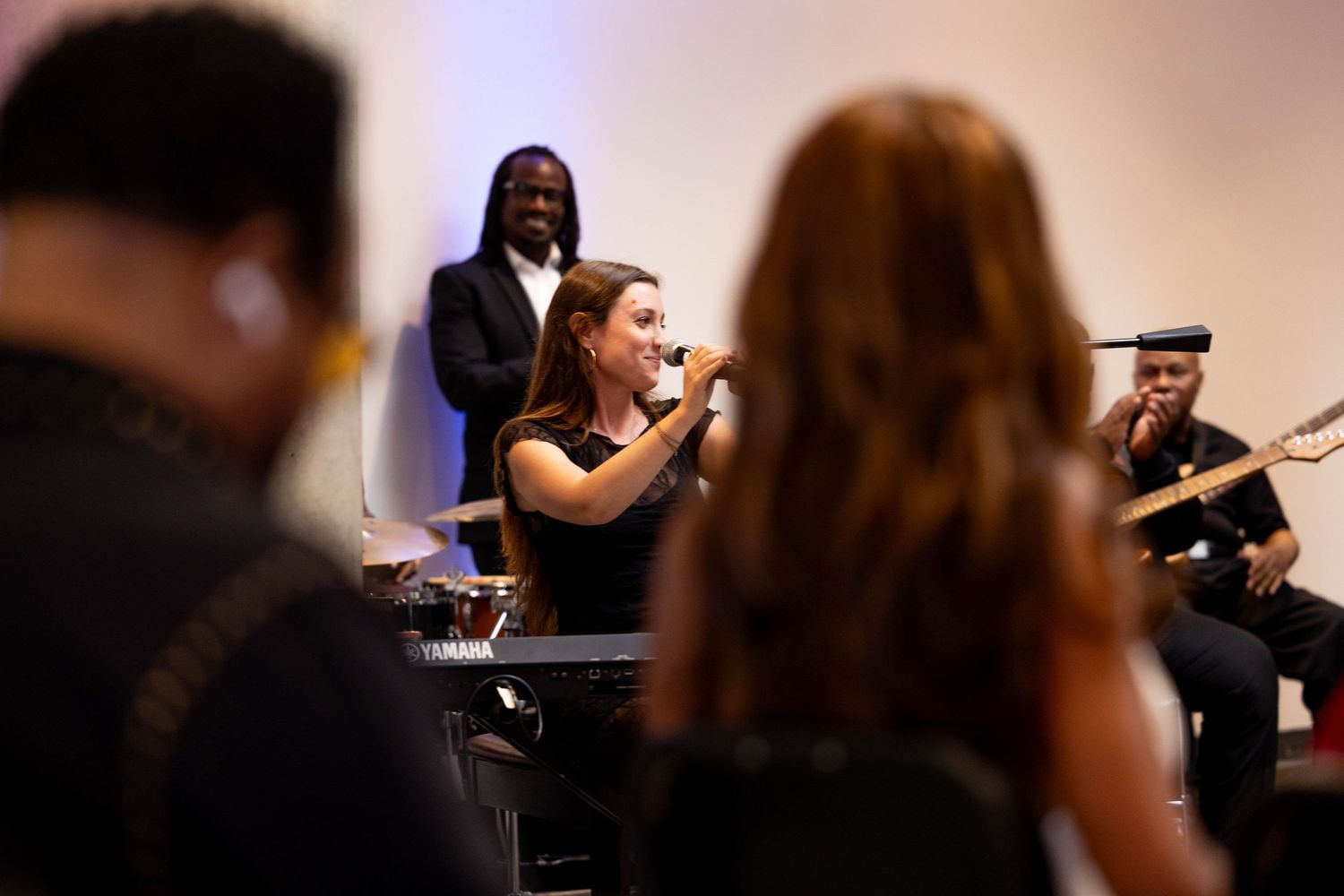 A female vocalist performs with a live band at an indoor event, smiling while holding a microphone near a Yamaha keyboard.