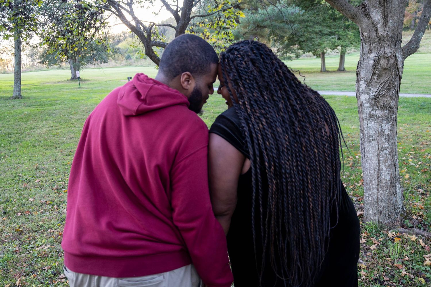 A couple shares an intimate forehead-to-forehead moment in a lush green park, the woman wearing long braided hair.