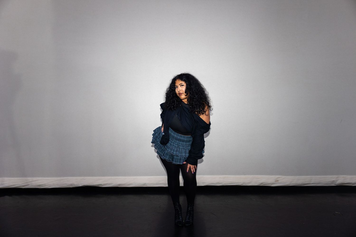 Woman in dark layered outfit with curly hair poses against gray wall.