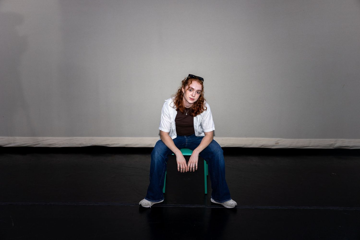 A young woman in jeans and white shirt sits casually on a black box on a dark studio floor.