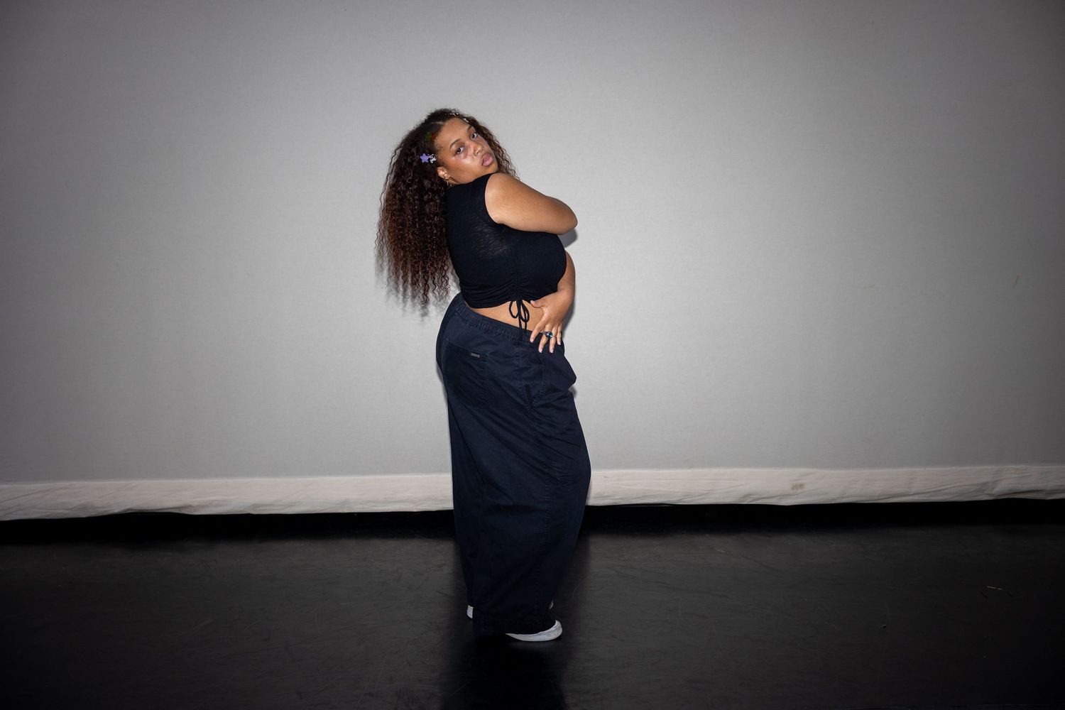 A curly-haired dancer poses in a navy crop top and wide-leg pants against a gray studio wall.