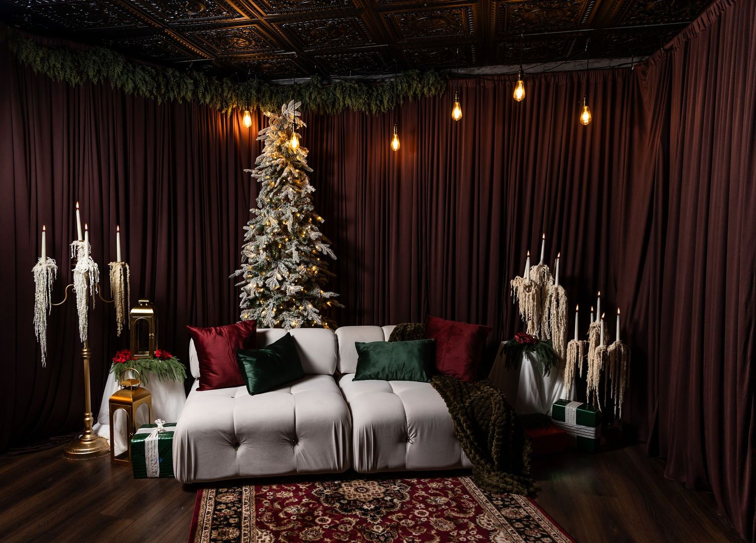 Cozy Christmas living room with a flocked tree, gray tufted sofa, red and green pillows, string lights, and burgundy velvet curtains.